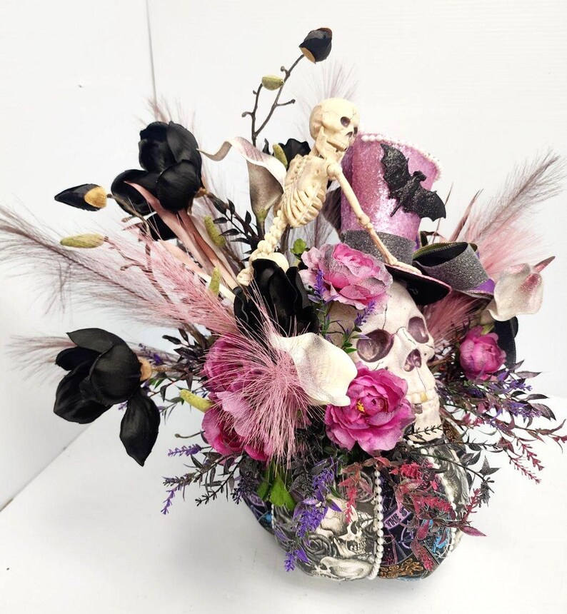 K&ouml;nnte beinhalten: Ein Halloween-Blumenarrangement mit einem Sch&auml;del mit Zylinder, einem Skelett, schwarzen und pinkfarbenen Blumen und verschiedenen Gr&uuml;npflanzen.