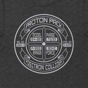 May include: A dark heather grey t-shirt with a white graphic of a proton pack from the movie Ghostbusters. The graphic includes the text "PROTON PACK", "POSITRON COLLIDER", "MODEL 84-89", "PHASE 7W33", "P-95G RANGE", "90HZ FORCE", "SRL NO G86-1", and "CTRL NO 3-62m".