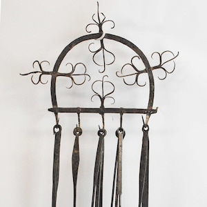 May include: An antique wrought iron kitchen utensil rack with six hanging tools. The rack has a decorative arched top with floral accents. The tools include a ladle, skimmer, spatula, and tongs, all with aged, rustic finishes.