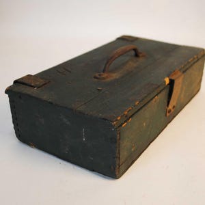 May include: A rectangular wooden storage box painted in a dark, weathered green finish. The box features a metal handle on the top lid, a metal latch on the front side, and visible signs of wear, including chipped paint and rusted metal hardware.