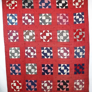 May include: A vintage quilt with a red background and a repeating pattern of square blocks. Each block features a central square surrounded by four triangles in various colors, including red, blue, white, and gray. The quilt is likely handmade.