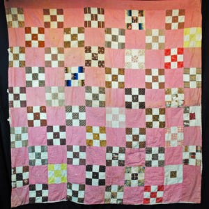 Puede incluir: Un edredón patchwork vintage con fondo rosa y patrón de tablero de ajedrez. Los cuadrados presentan varias telas en tonos marrón, blanco y rojo. El edredón está colgado, mostrando su construcción y diseño hechos a mano.