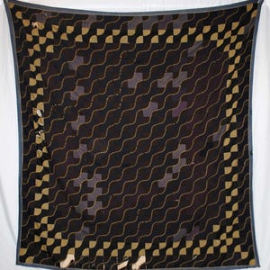 Op de afbeelding: Een vierkante, zwarte quilt met een herhalend patroon van golvende lijnen en vierkanten in goud en paars. De quilt heeft een donkerblauwe rand en wordt gepresenteerd op een witte achtergrond.