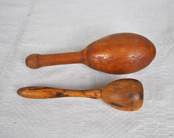 May include: Two wooden maracas, one larger than the other, with a smooth, polished finish. The maracas are a warm brown color and appear to be handmade. They are lying on a white, textured surface.