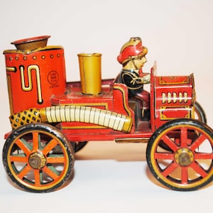 May include: A vintage red and gold toy fire engine with a firefighter figure. The toy has black wheels with red and gold detailing. The words "Lin Mar Toys Japan" are visible on the side.