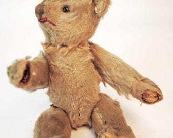 Puede incluir: Un pequeño oso de peluche de color marrón claro con pelaje texturizado y desgastado. El oso presenta una nariz oscura cosida, una boca cosida en rojo y un desgaste visible en las almohadillas de las patas y extremidades. Está sentado con un brazo extendido.