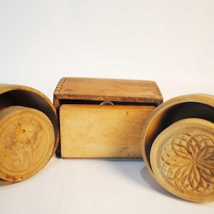 Peut inclure: Trois ustensiles de cuisine anciens en bois. Deux sont des moules à beurre ronds avec des motifs sculptés et un est une boîte rectangulaire en bois. Les articles sont en bois de couleur claire et reposent sur un fond blanc.