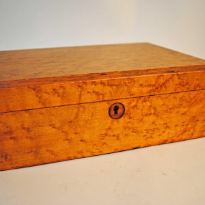 May include: A rectangular wooden box featuring a light brown burl wood grain pattern throughout the exterior. The box has a hinged lid and a small, centered keyhole on the front panel. The wood displays a smooth finish with dark edges.