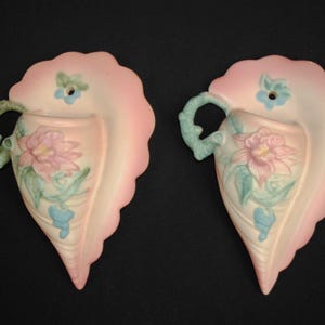 May include: Two vintage wall pocket vases. Each vase is leaf-shaped with a scalloped edge, painted in pink and cream. They feature a floral design with pink and blue accents and a green handle. The vases are approximately 8 inches tall.
