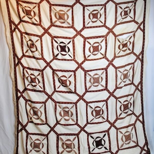 May include: Cream-coloured quilt with a geometric pattern. The quilt features a repeating design of squares and octagons in a deep brown colour. The squares contain a smaller, patterned design. The quilt is likely made of cotton or a similar fabric.
