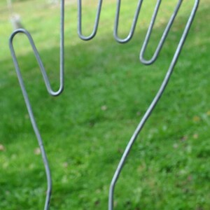Może przedstawiać: Metalowa rzeźba dłoni z szarym wykończeniem. Rzeźba wykonana jest z giętych metalowych pręt&oacute;w, tworzących kształt otwartej dłoni. Jest umieszczona na zewnątrz na zielonym trawniku.