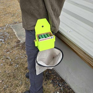 May include: A neon green shotgun shell holder with a white bag attached. The holder is labeled "WINCHESTER" and contains green shotgun shells. The bag is made of white fabric with a black trim.