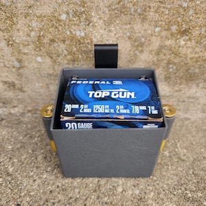 May include: A gray plastic 20-gauge ammunition carrier with gold-colored accents. The carrier holds a box of Federal Top Gun shotgun shells. The box is blue with white text, including "20 GAUGE" and other specifications.