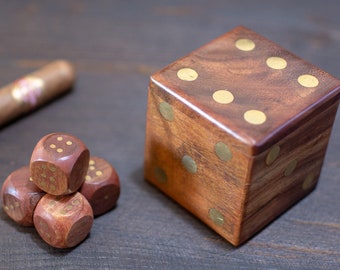 May include: A wooden dice set and a cigar on a dark wood surface. The set includes a large, square box-shaped die and four smaller dice. The dice have gold-coloured dots. The cigar is brown with a pink and gold label.