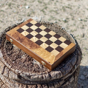 May include: A wooden chessboard with a brown and light wood checkered pattern. The board is closed and sitting on a tree stump.