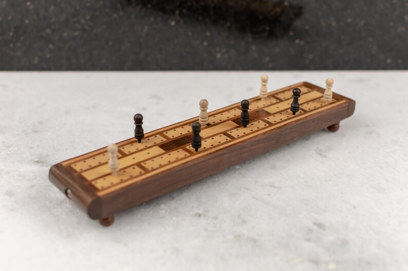 May include: A wooden cribbage board with black and white pegs. The board is made of walnut wood and has a light brown finish. The pegs are arranged in a pattern on the board.