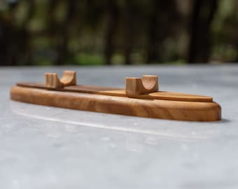 May include: A wooden pen holder with a rectangular base and two curved supports. The pen holder is made of light brown wood and has a smooth finish. The supports are designed to hold pens or pencils upright.