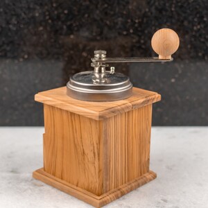 May include: A wooden manual coffee grinder with a silver metal top and a wooden crank handle. The grinder is sitting on a white surface.