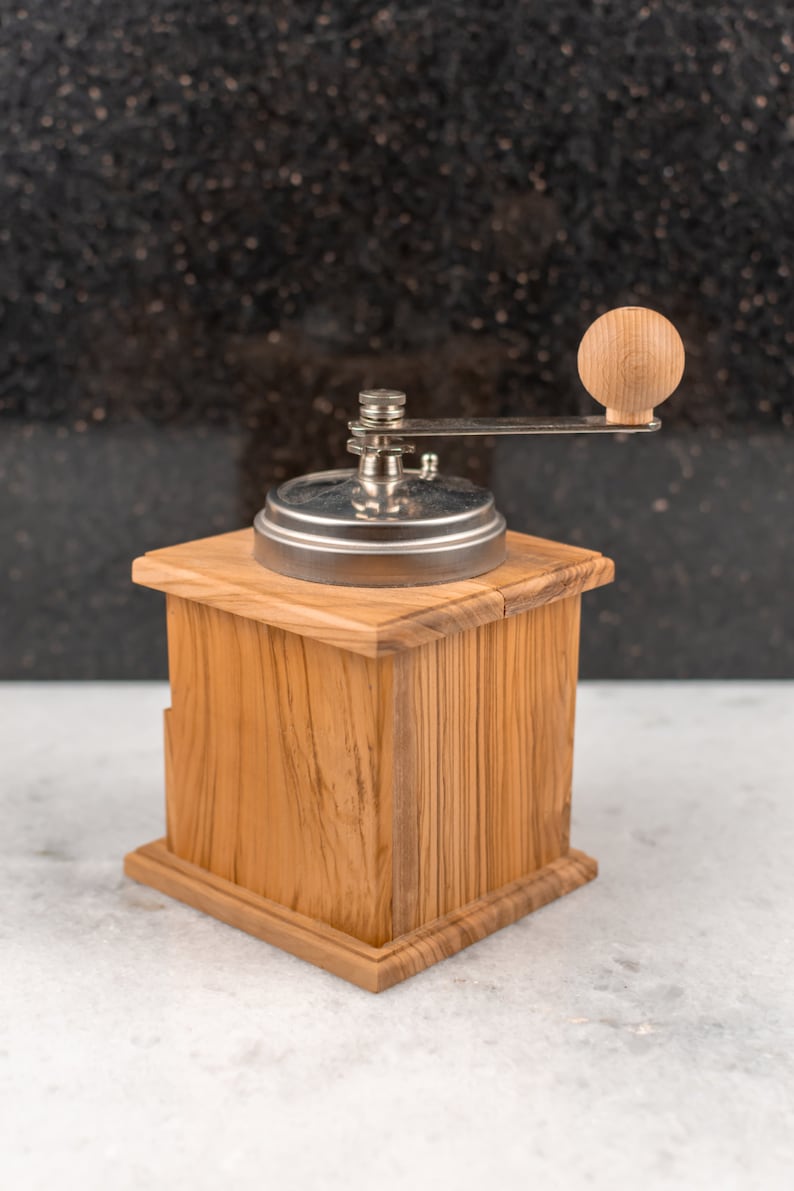 May include: A wooden manual coffee grinder with a silver metal top and a wooden crank handle. The grinder is sitting on a white surface.