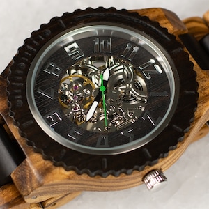 Puede incluir: Un reloj de pulsera de madera con una banda de madera marrón y negra y una esfera plateada con un movimiento esqueletizado. El reloj tiene una aguja de segundos verde.