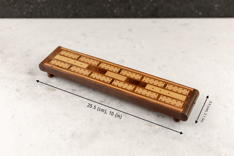 May include: A wooden cribbage board with a light brown and dark brown wood inlay design. The board measures 10 inches long and 2.1 inches wide.