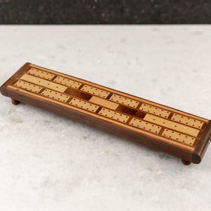 May include: A wooden cribbage board with a light brown and dark brown wood inlay design. The board has a rectangular shape and is about 12 inches long.