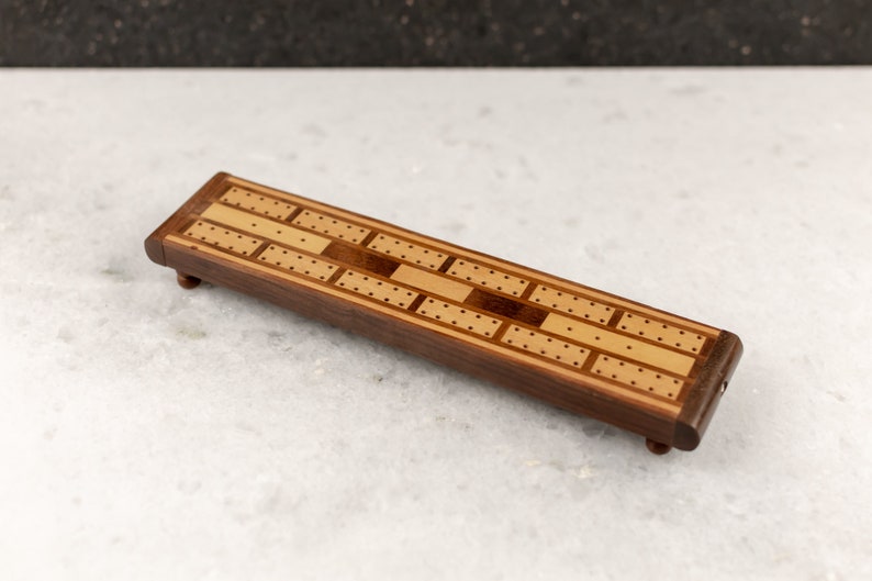 May include: A wooden cribbage board with a light brown and dark brown wood inlay design. The board has a rectangular shape and is about 12 inches long.
