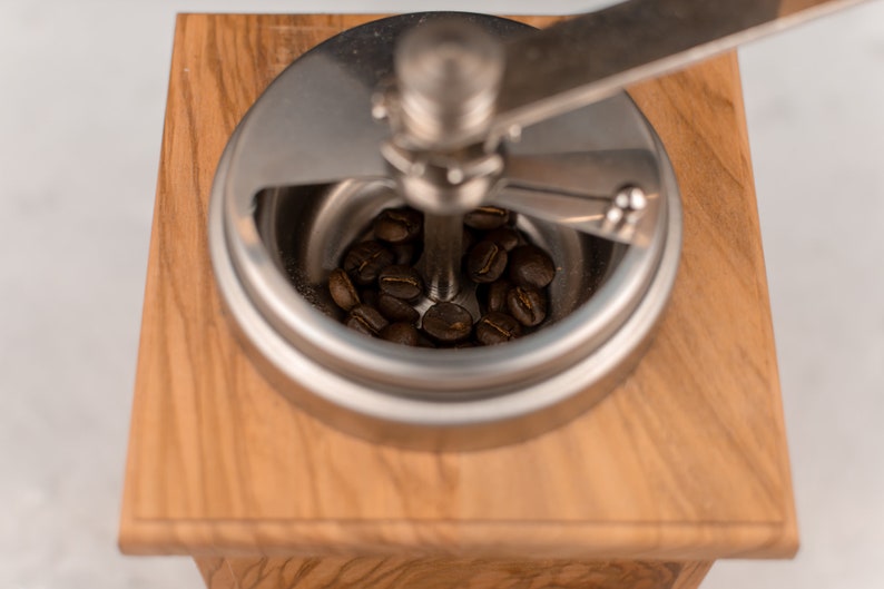 May include: A close-up of a wooden coffee grinder with a metal top. The grinder is filled with coffee beans.