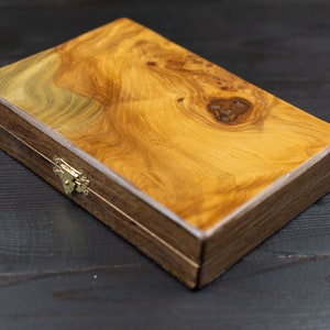 May include: A wooden box with a natural wood grain finish and a brass latch. The box is closed and sitting on a dark wood surface.