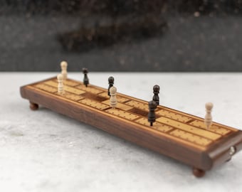 May include: A wooden cribbage board with black and white pegs. The board is made of walnut wood and has a light brown finish. The pegs are arranged in a line down the center of the board.