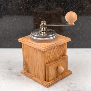 May include: A wooden coffee grinder with a metal crank and a drawer. The grinder is made of light brown wood with a natural grain pattern. The crank is silver and has a wooden knob. The drawer is also made of wood and has a small wooden knob.