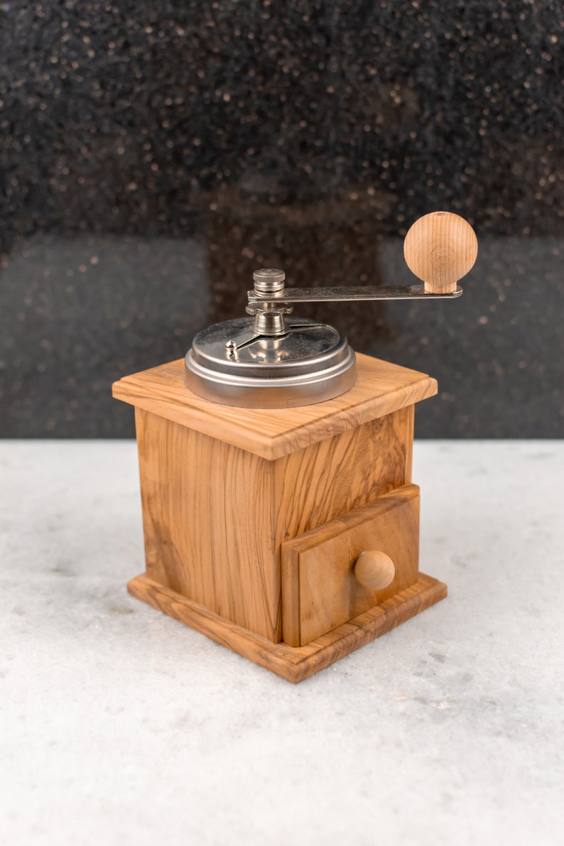 May include: A wooden coffee grinder with a metal crank and a drawer. The grinder is made of light brown wood with a natural grain pattern. The crank is silver and has a wooden knob. The drawer is also made of wood and has a small wooden knob.