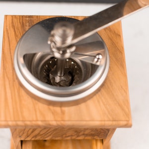 May include: A wooden manual coffee grinder with a silver crank and a silver grinding mechanism. The grinder is made of light wood with a grain pattern.