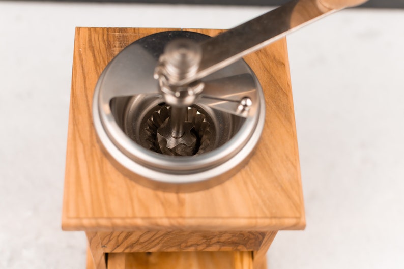 May include: A wooden manual coffee grinder with a silver crank and a silver grinding mechanism. The grinder is made of light wood with a grain pattern.