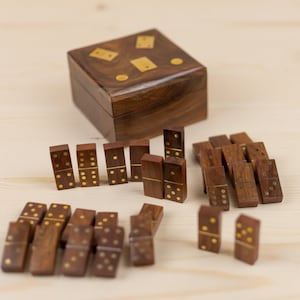 May include: A wooden domino set with a matching box. The dominoes are dark brown with gold-colored dots. The box is square and has gold-colored accents on the lid. The set is displayed on a light wood surface.
