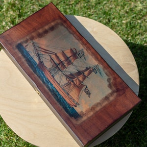 May include: A wooden box with a hinged lid, featuring a vintage illustration of a sailing ship with billowing sails against a blue ocean background. The box has a dark brown finish and a brass latch.
