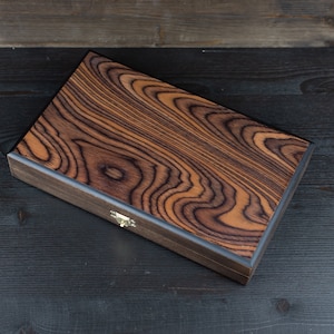 May include: A wooden box with a dark brown wood grain pattern and a brass latch. The box is closed and sitting on a dark wood surface.