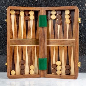 Puede incluir: Juego de backgammon de madera abierto con triángulos marrones claros y oscuros, fieltro verde y fichas de madera. El juego está abierto, revelando la superficie de juego y las piezas. La caja de madera tiene un cierre dorado.