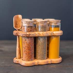 May include: A wooden spice rack with six glass jars filled with different spices. The jars have cork tops and are arranged in a circle. A wooden spoon is included.