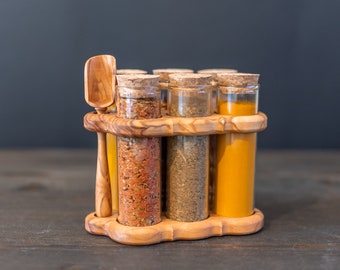May include: A wooden spice rack with six glass jars filled with different spices. The jars have cork tops and are arranged in a circle. A wooden spoon is included.