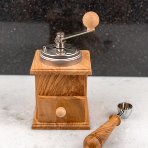May include: A wooden coffee grinder with a metal crank and a drawer. A wooden scoop with a metal top is next to the grinder.