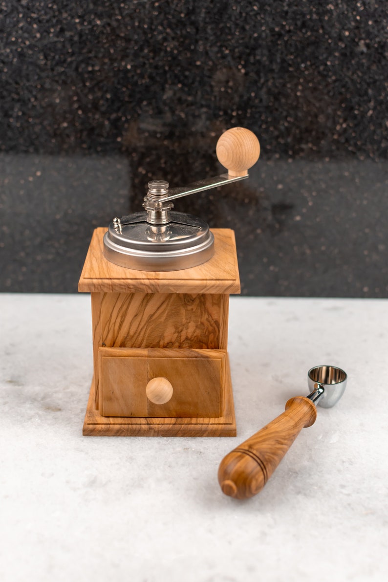 May include: A wooden coffee grinder with a metal crank and a drawer. A wooden scoop with a metal top is next to the grinder.