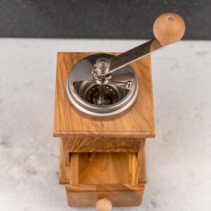 May include: A wooden coffee grinder with a metal crank and a drawer for storing coffee beans. The grinder is made of light brown wood and has a natural finish.