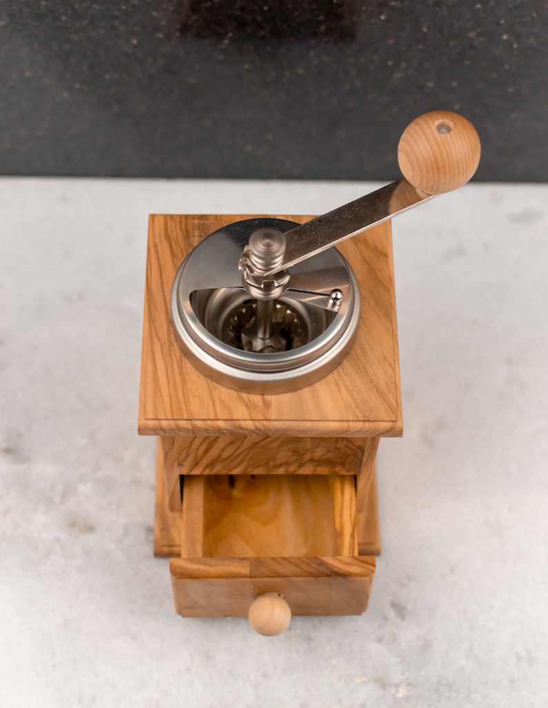 May include: A wooden coffee grinder with a metal crank and a drawer for storing coffee beans. The grinder is made of light brown wood and has a natural finish.