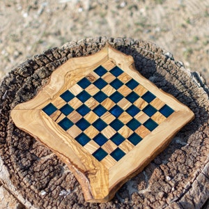 May include: A handcrafted wooden chess board with a unique design. The board features a natural wood frame with a square playing surface of alternating light wood and dark blue squares. The board is set on a tree stump.