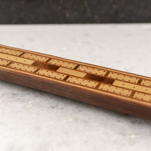 May include: A wooden cribbage board with a light brown and dark brown inlay design. The board has small pegs for scoring and is made of walnut wood.