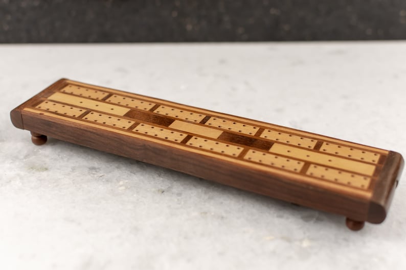 May include: A wooden cribbage board with a light brown and dark brown inlay design. The board has small pegs for scoring and is made of walnut wood.