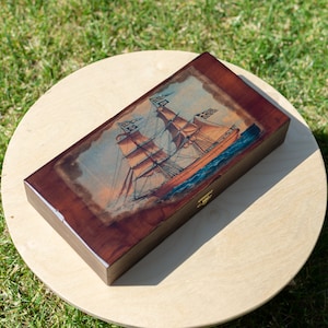 May include: A wooden box with a hinged lid, featuring a vintage illustration of a sailing ship with billowing sails against a blue sky and ocean waves. The box has a dark brown wood finish and a brass latch.