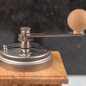 May include: A close-up of a wooden coffee grinder with a silver metal top and a wooden knob. The grinder is on a wooden base.