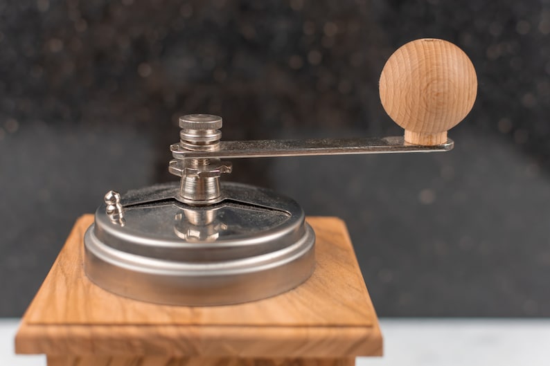 May include: A close-up of a wooden coffee grinder with a silver metal top and a wooden knob. The grinder is on a wooden base.